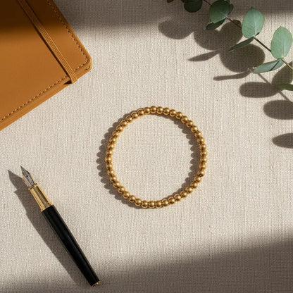 Flat-lay of a gold beaded stretch bracelet on a textured cream fabric surface beside a mustard notebook, fountain pen, and eucalyptus leaves, styled for a chic desk look | Oria Jewel