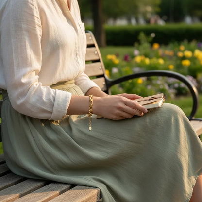 Woman relaxing on a park bench at golden hour, holding a book on her lap while the gold chain link bracelet glows softly against her wrist, framed by a flowing skirt and garden flowers in the background | Oria Jewel