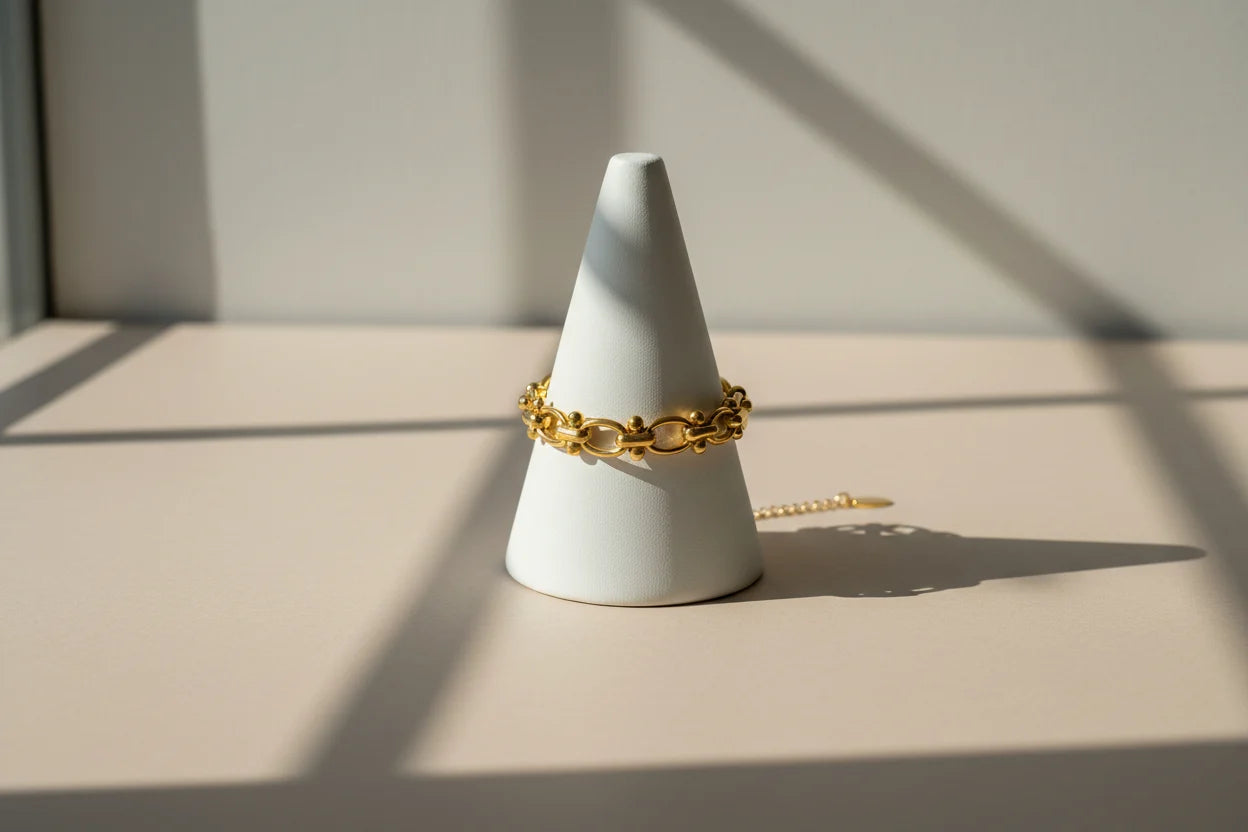 Minimalist product display of a gold chain link bracelet wrapped around a white cone stand, with sharp window shadows creating a modern, gallery-style look | Oria Jewel