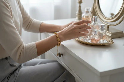 Woman seated at a dressing table, reaching for perfume bottles while a gold chain link bracelet hangs gracefully from her wrist, completing her soft neutral outfit | Oria Jewel