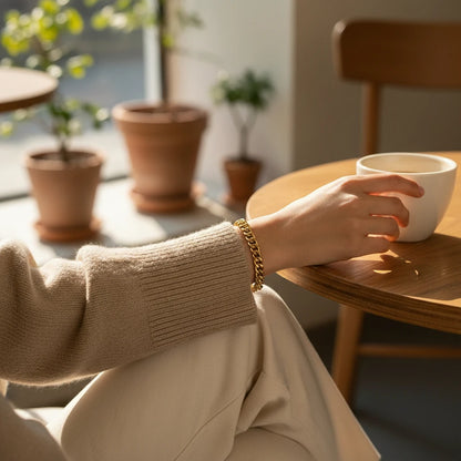 Cozy café close-up of a wrist wearing a gold curb chain bracelet styled with neutral knit sweater and coffee cup | Oria Jewel