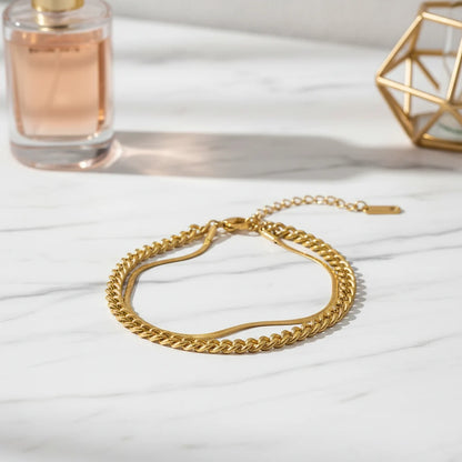 Minimalist gold double chain bracelet resting on white marble beside perfume bottle, softly lit for everyday elegance | Oria Jewel
