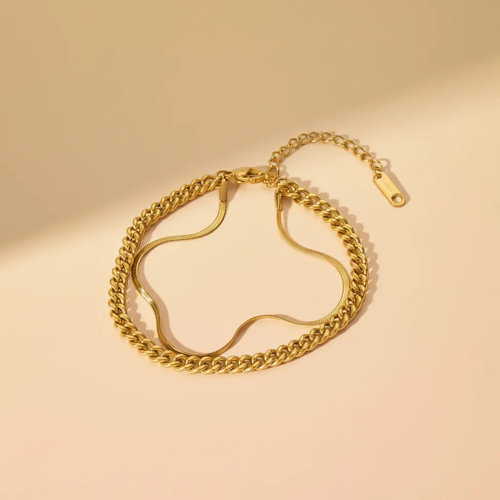 Delicate gold double chain bracelet displayed on warm beige surface with soft shadows, highlighting its layered shine | Oria Jewel