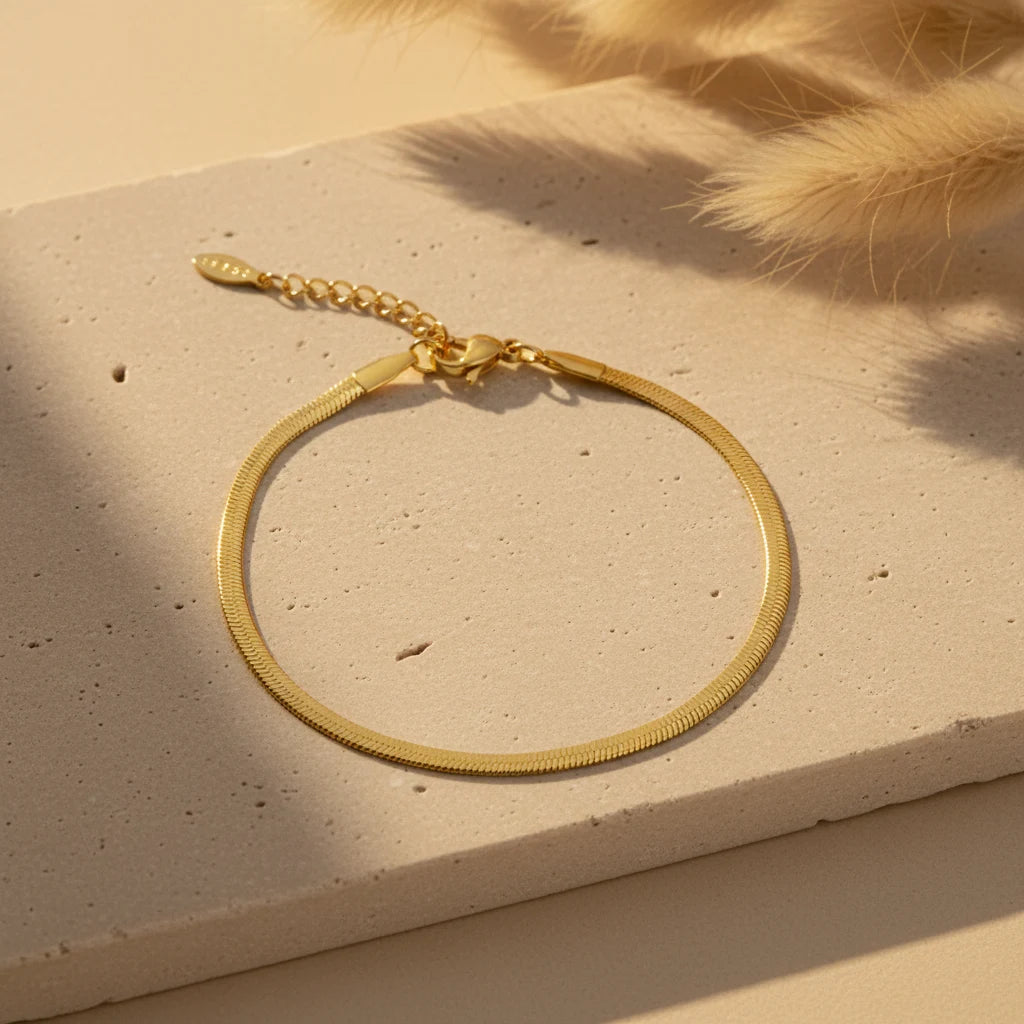 Gold herringbone chain bracelet laid in a circle on a textured beige stone surface with warm sunlight and soft pampas grass in the corner | Oria Jewel