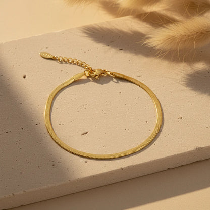 Gold herringbone chain bracelet laid in a circle on a textured beige stone surface with warm sunlight and soft pampas grass in the corner | Oria Jewel