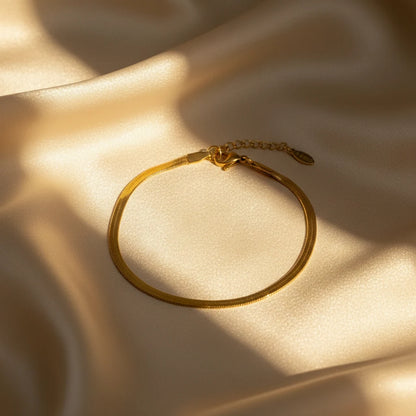 Elegant gold herringbone chain bracelet arranged in a circle on softly rippled champagne satin fabric with warm sunlight and shadows | Oria Jewel
