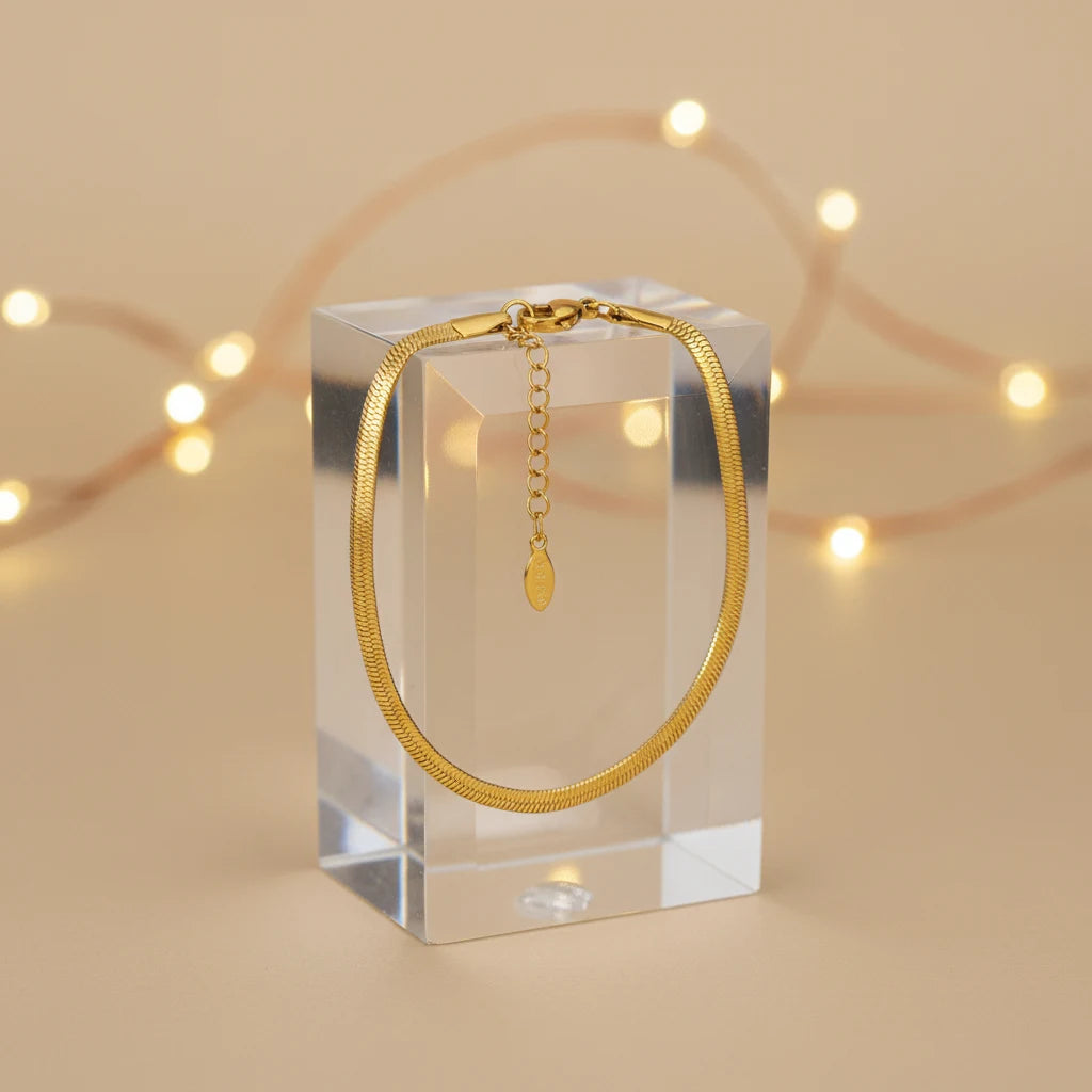 Gold herringbone chain bracelet hanging neatly over a clear acrylic block with soft fairy lights in the background for a festive look | Oria Jewel