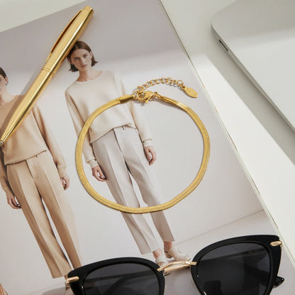 Fashion flat lay of a gold herringbone chain bracelet on a magazine page with minimalist outfits, next to sunglasses and a gold pen | Oria Jewel