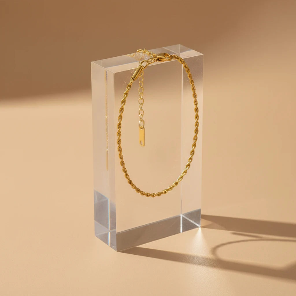Gold rope chain bracelet hanging straight from a clear acrylic block on a warm beige background, casting a soft shadow for modern display | Oria Jewel
