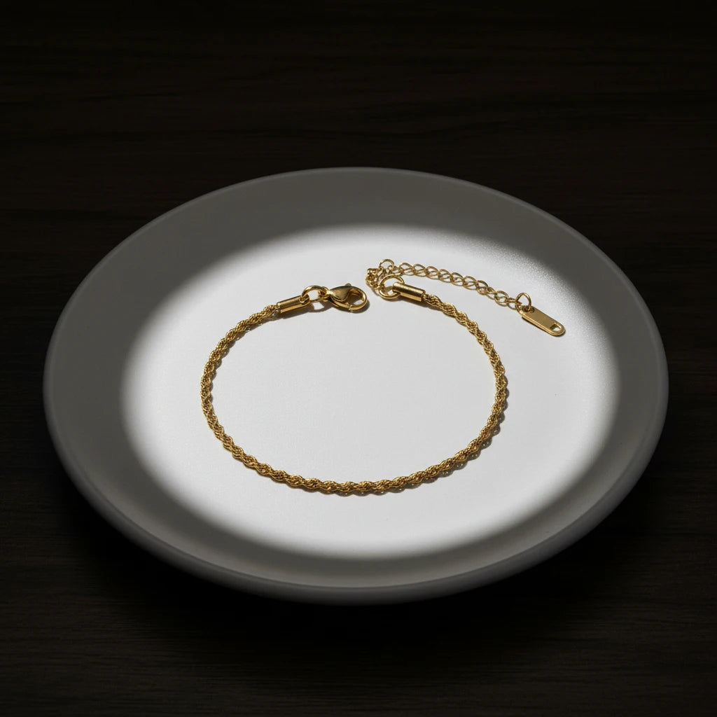 Dramatic spotlight image of a gold rope chain bracelet centered on a round white dish against a dark background for high-contrast presentation | Oria Jewel