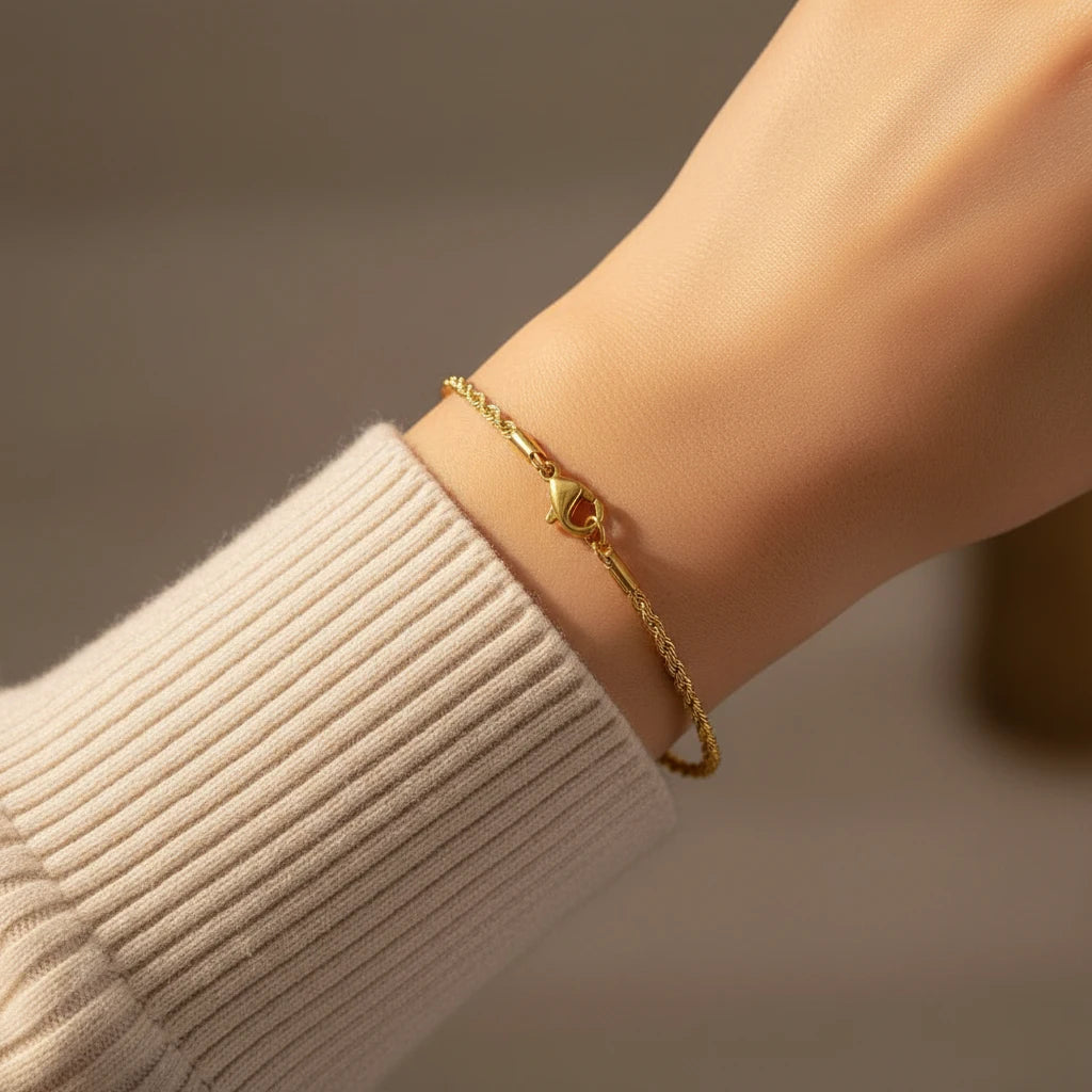 Close-up of a wrist in a ribbed beige sweater sleeve showing the clasp side of a dainty gold rope chain bracelet, softly lit and minimal | Oria Jewel