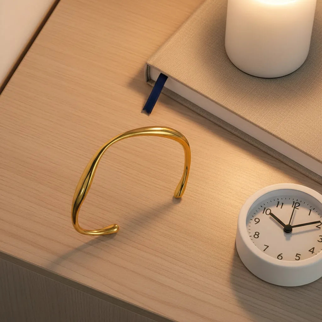 Minimalist gold cuff bracelet resting on a wooden bedside table beside a lit candle, closed notebook and small white clock, softly lit evening scene | Oria Jewel