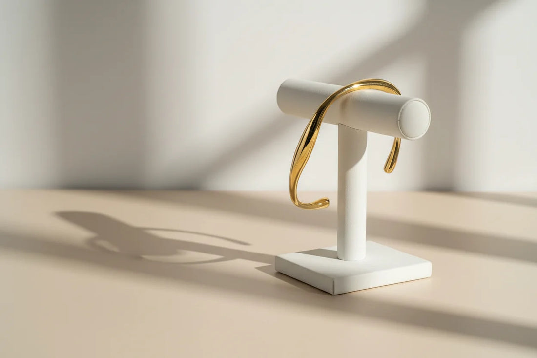Minimalist gold cuff bracelet draped over a white jewelry stand, casting a soft shadow in warm natural window light on a beige surface | Oria Jewel