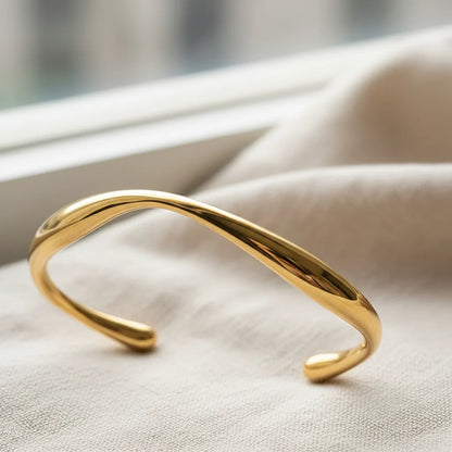 Close-up of minimalist gold cuff bracelet lying on light beige fabric near a window, polished surface reflecting soft daylight and emphasizing the fluid curve | Oria Jewel