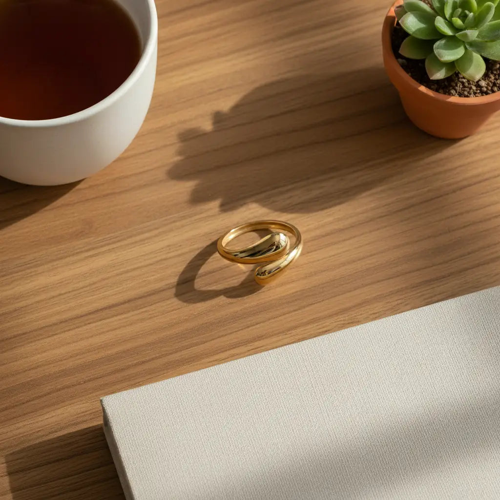 open gold wrap ring on a warm wooden desk beside a white cup of tea and a small succulent, with sunlit shadows highlighting the smooth bypass shape | OriaJewel