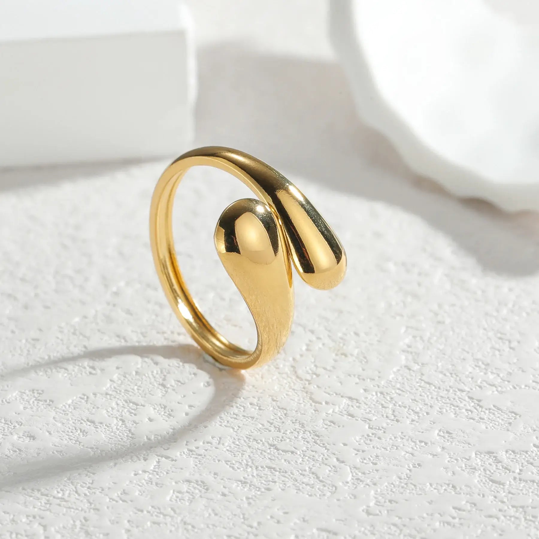 close-up of an open gold wrap ring standing upright on a textured white surface, bold sculpted ends and bright highlights for a premium look | OriaJewel