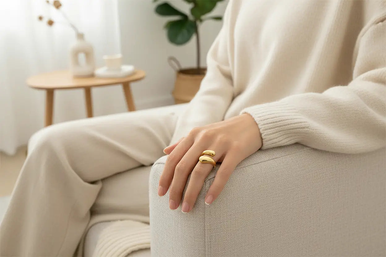 lifestyle shot of an open gold wrap ring worn on a hand resting on a sofa arm, cozy neutral knit outfit and soft daylight for everyday styling | OriaJewel