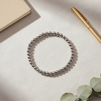 Flat-lay image of a Silver Beaded Stretch Bracelet arranged in a circle on a textured cream fabric surface, styled next to a beige notebook, gold pen and eucalyptus leaves for a chic desk look | Oria Jewel