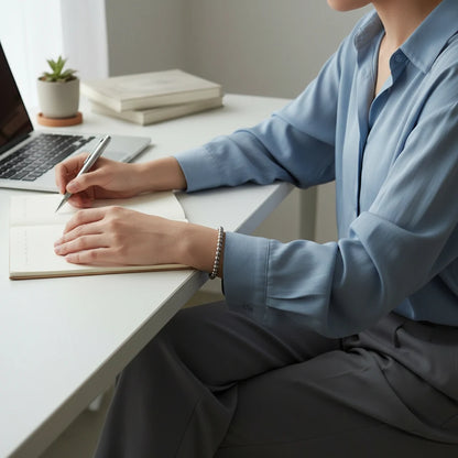 Woman working at a clean white desk, writing in a notebook next to a laptop while wearing a subtle Silver Beaded Stretch Bracelet that adds a polished touch to her office outfit | Oria Jewel