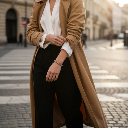 Street-style scene of a woman in a camel trench coat, white blouse and black trousers crossing a city crosswalk with a Silver Beaded Stretch Bracelet accenting her wrist in the evening light | Oria Jewel