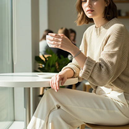Casual café moment with a woman in a beige knit sweater and cream trousers holding a cup of coffee, the Silver Beaded Stretch Bracelet providing a simple, modern sparkle on her wrist | Oria Jewel