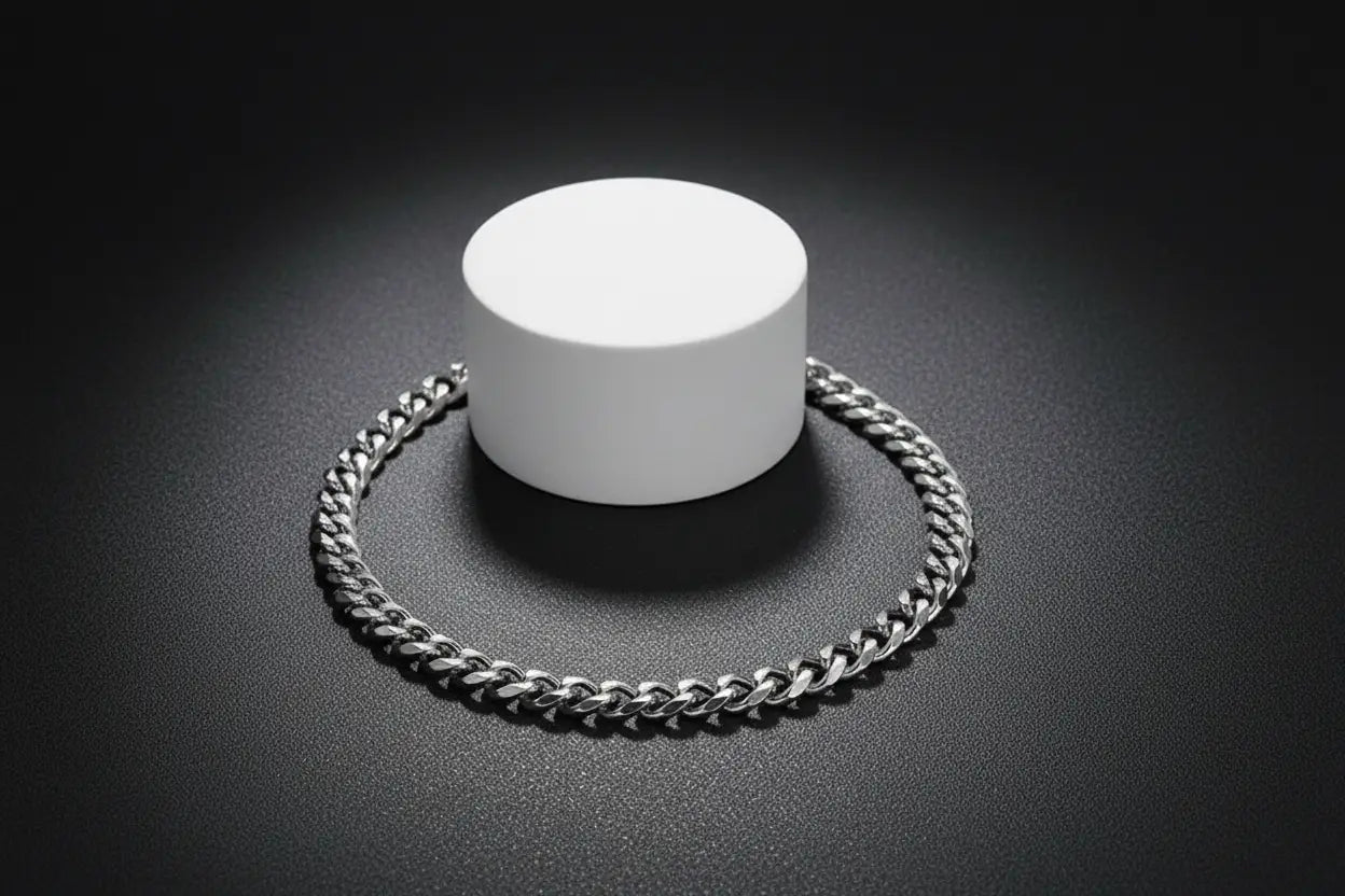 Close-up product shot of a sleek silver chain bracelet arranged in a circle around a white cylindrical stand, highlighted by dramatic lighting on a dark textured background | Oria Jewel