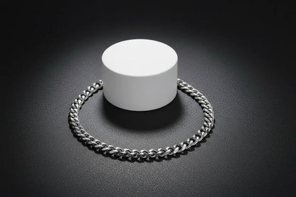 Close-up product shot of a sleek silver chain bracelet arranged in a circle around a white cylindrical stand, highlighted by dramatic lighting on a dark textured background | Oria Jewel