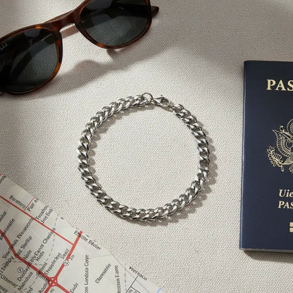 Flat lay of a travel scene featuring a shiny silver chain bracelet laid in a circle beside sunglasses, a navy blue passport, and a city map on a neutral fabric surface | Oria Jewel