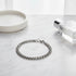 Elegant silver chain bracelet displayed in a circle on a white marble surface, styled with a minimal round dish and a clear glass perfume bottle for a clean, modern look | Oria Jewel