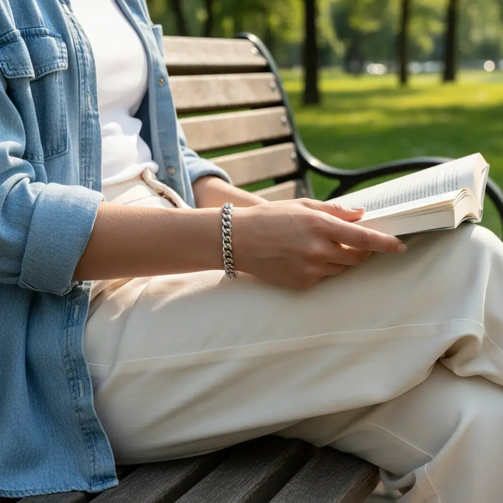 Casual outdoor lifestyle shot of a woman reading on a park bench, wearing a polished silver chain bracelet on her wrist, styled with a white top and light pants under a denim shirt | Oria Jewel