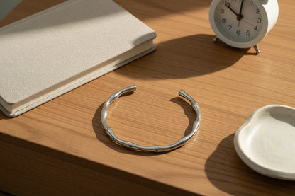 Bamboo-style silver cuff bracelet lying open on a wooden bedside table beside a fabric notebook, ceramic trinket dish and alarm clock in warm sunlight | Oria Jewel