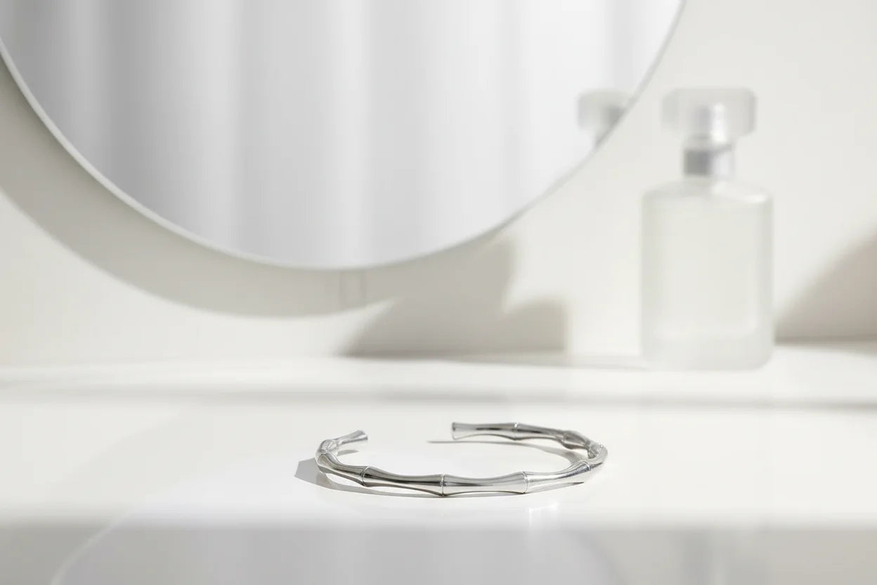 Minimal bathroom vanity scene with a slim silver cuff bracelet placed on a glossy white counter in front of a round mirror and frosted perfume bottle | Oria Jewel