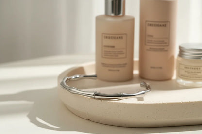 Self-care vignette with a polished silver cuff bracelet resting on a round ceramic tray beside neutral-toned skincare bottles in soft diffused daylight | Oria Jewel