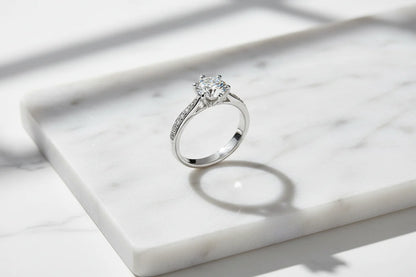 Clean marble flat-lay featuring a silver cz solitaire ring standing upright with crisp natural light, a defined shadow ring, and a bright sparkle on the round CZ stone | OriaJewel
