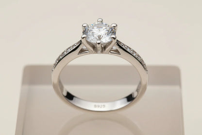 Macro front view of a silver cz solitaire ring on a clear acrylic display block, highlighting the round CZ centerpiece, pavé side stones, and the 925 marking inside the band | OriaJewel