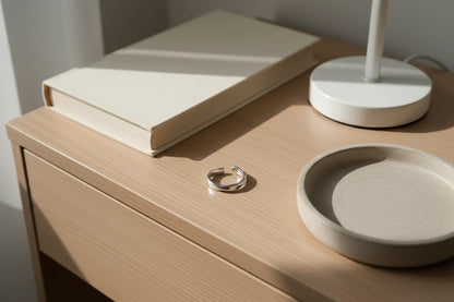 Silver wave ring resting on a light wooden bedside table beside a neutral book, white lamp base, and ceramic tray, with warm sunlight and soft shadows | OriaJewel