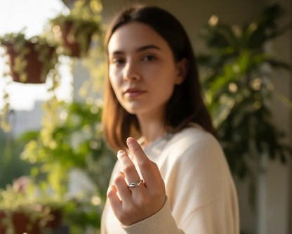 Lifestyle shot of a woman holding her hand forward while wearing a silver wave ring, soft golden-hour light with leafy balcony background and natural bokeh | OriaJewel