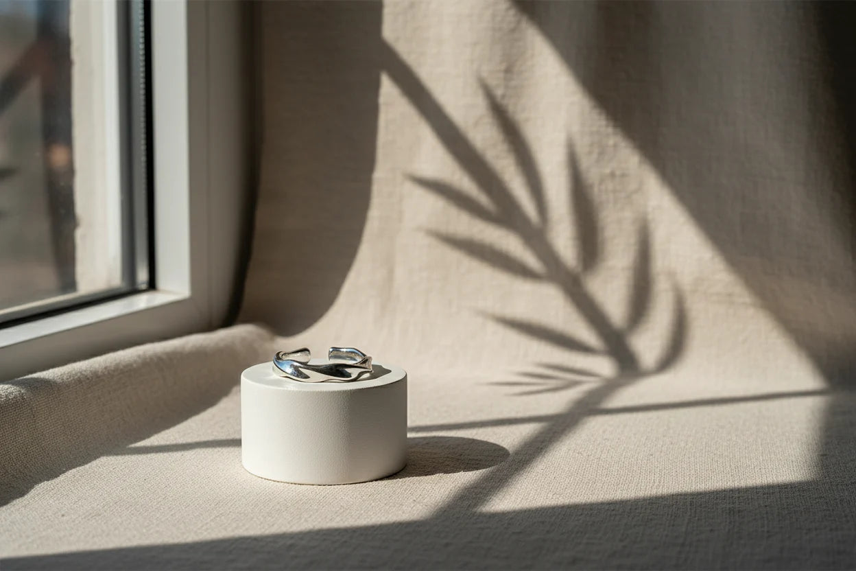 Product-only scene of a silver wave ring on a small white cylindrical stand near a window, dramatic plant-shadow pattern across neutral fabric | OriaJewel