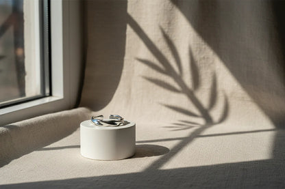 Product-only scene of a silver wave ring on a small white cylindrical stand near a window, dramatic plant-shadow pattern across neutral fabric | OriaJewel