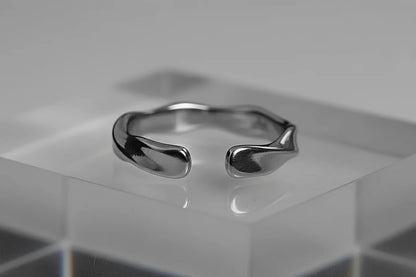Studio close-up of a silver wave ring on a clear acrylic block, showing the open-front wave ends and high-polish reflective finish against a gray backdrop | OriaJewel