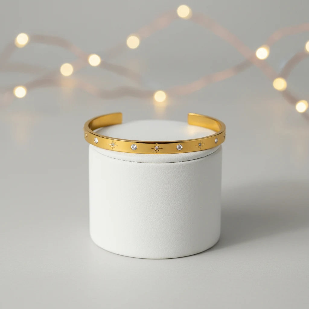 Gold star cuff bracelet displayed on a white cylindrical stand with soft fairy lights in the background, emphasizing its delicate sparkle and open-ended design | Oria Jewel