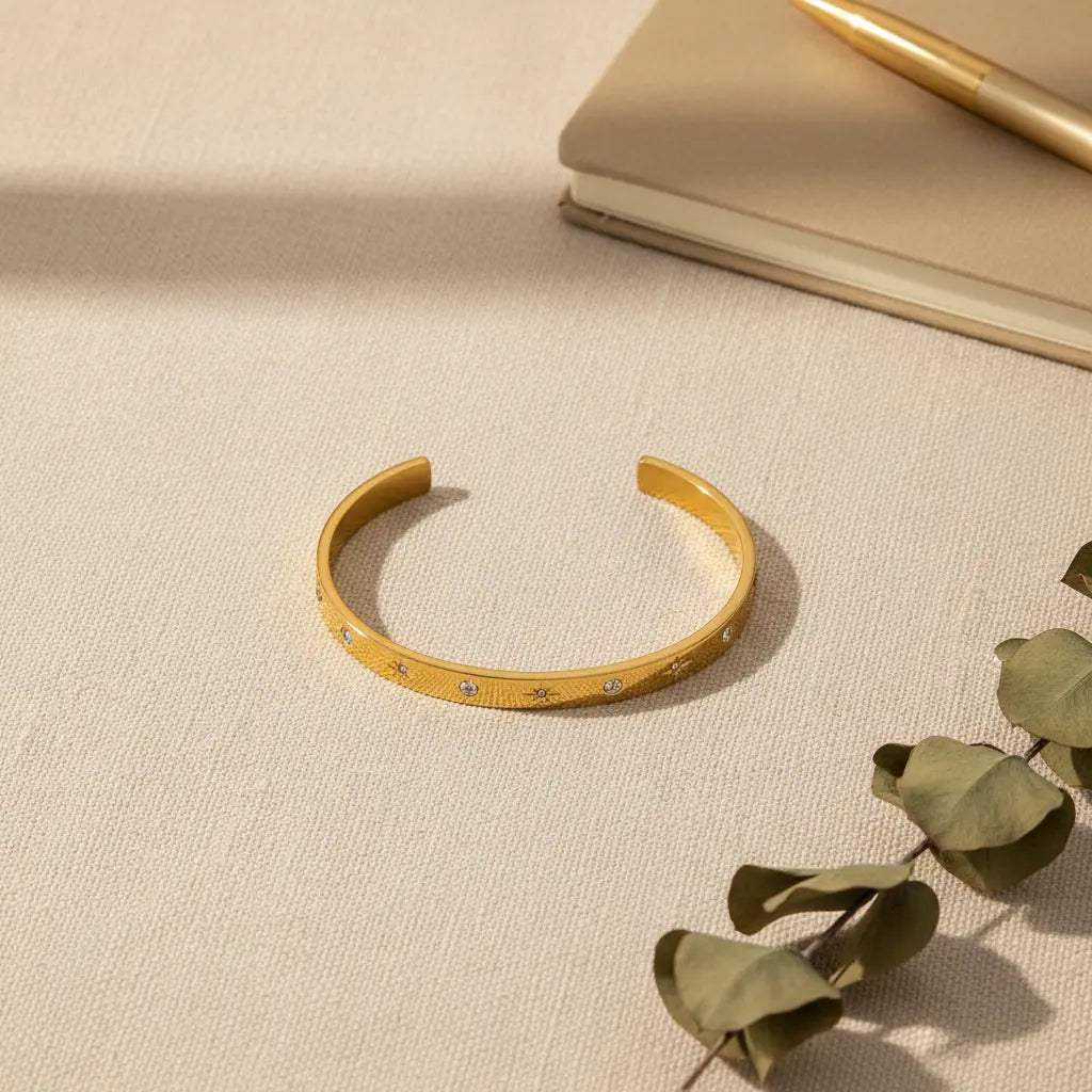 Flat-lay of a gold star cuff bracelet on textured beige fabric next to a neutral notebook, gold pen and eucalyptus leaves, styled as a chic desk-side accessory | Oria Jewel
