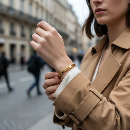 Woman in a camel trench coat adjusting a sleek gold statement cuff bracelet on her wrist while walking through a city street, highlighting its modern everyday style | Oria Jewel