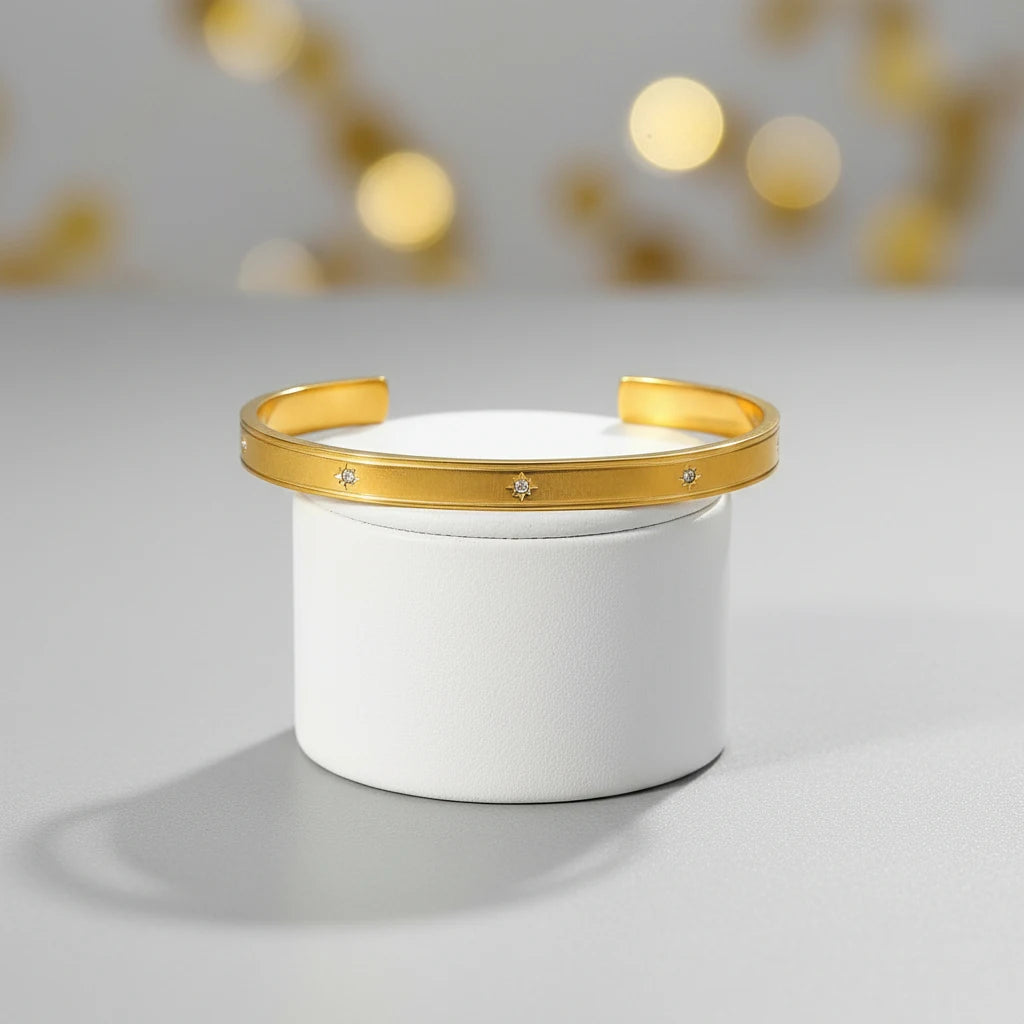 Studio shot of a gold statement cuff bracelet balanced on a white cylindrical stand with soft golden bokeh in the background, emphasizing its clean lines and subtle sparkle | Oria Jewel