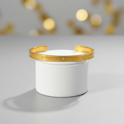 Studio shot of a gold statement cuff bracelet balanced on a white cylindrical stand with soft golden bokeh in the background, emphasizing its clean lines and subtle sparkle | Oria Jewel