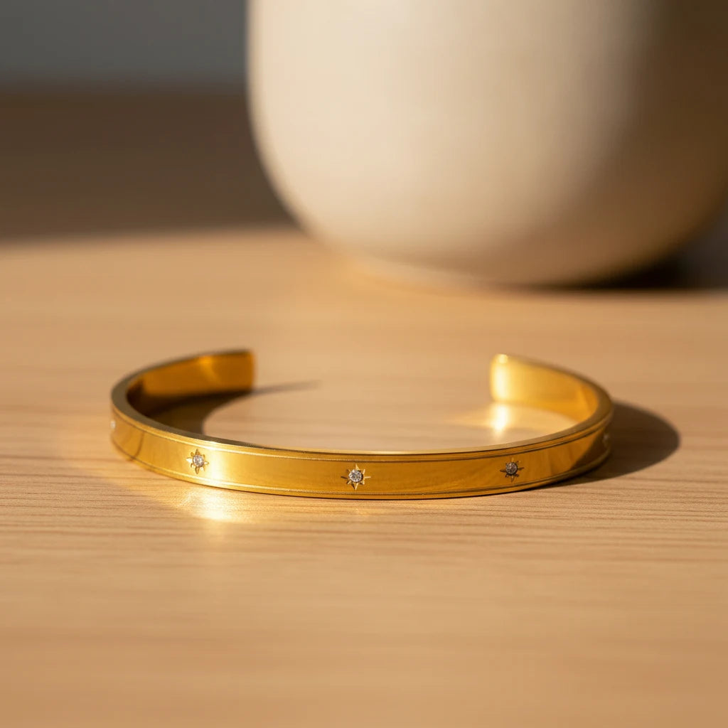 Minimalist scene of a gold statement cuff bracelet resting open on a light wooden surface beside a neutral ceramic vase, bathed in warm natural sunlight | Oria Jewel