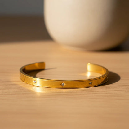 Minimalist scene of a gold statement cuff bracelet resting open on a light wooden surface beside a neutral ceramic vase, bathed in warm natural sunlight | Oria Jewel