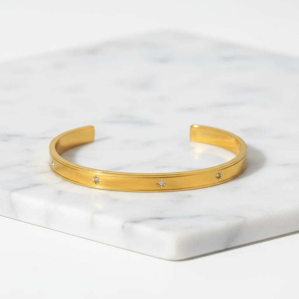 Gold statement cuff bracelet displayed diagonally on a white marble slab, its slim band dotted with small sparkling stones for a refined, luxe finish | Oria Jewel