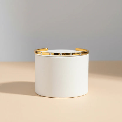 Studio product shot of a thin gold cuff bracelet standing upright around a white cylindrical display on a beige surface, highlighting its minimalist open-ended design and crystal accents | Oria Jewel
