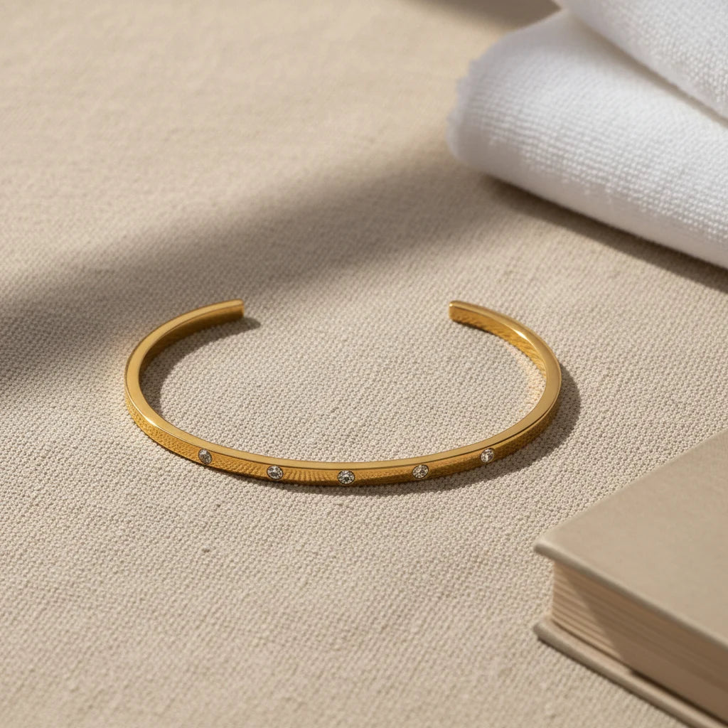 Thin gold cuff bracelet displayed on a neutral fabric surface beside folded white towels and a beige book, styled to suggest an elegant, everyday jewelry essential | Oria Jewel